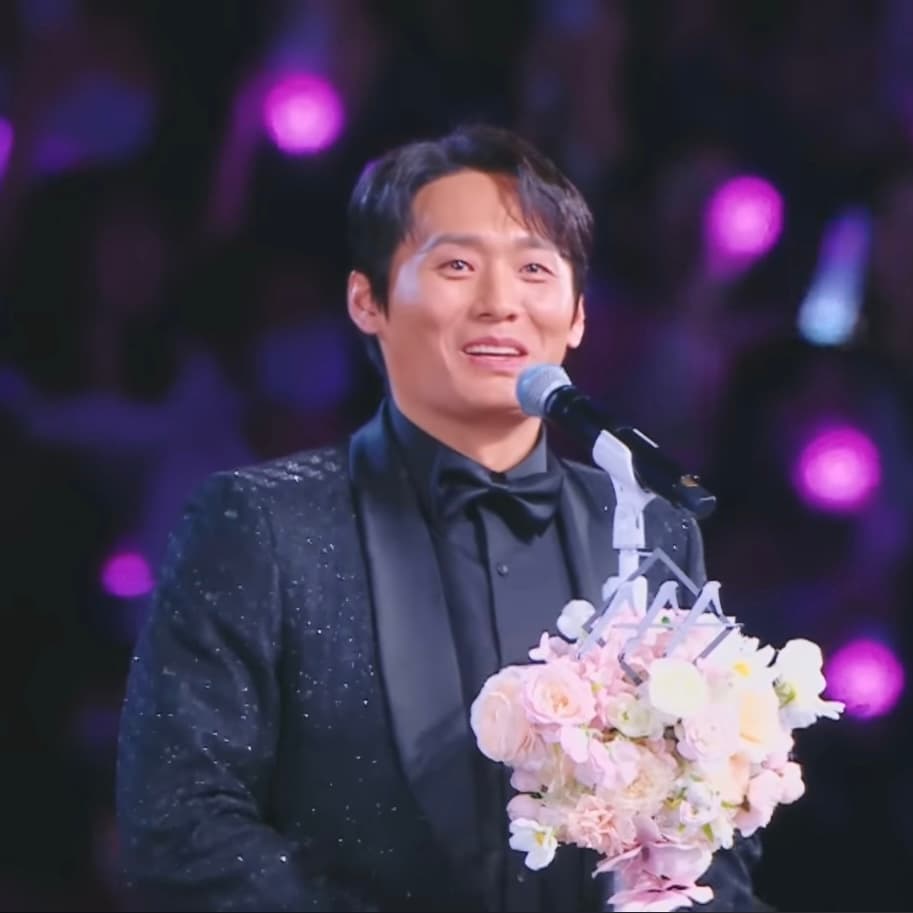 Choi Dae-hoon in a black suit speaking into a microphone and holding flowers at the 2025 Asia Artist Awards