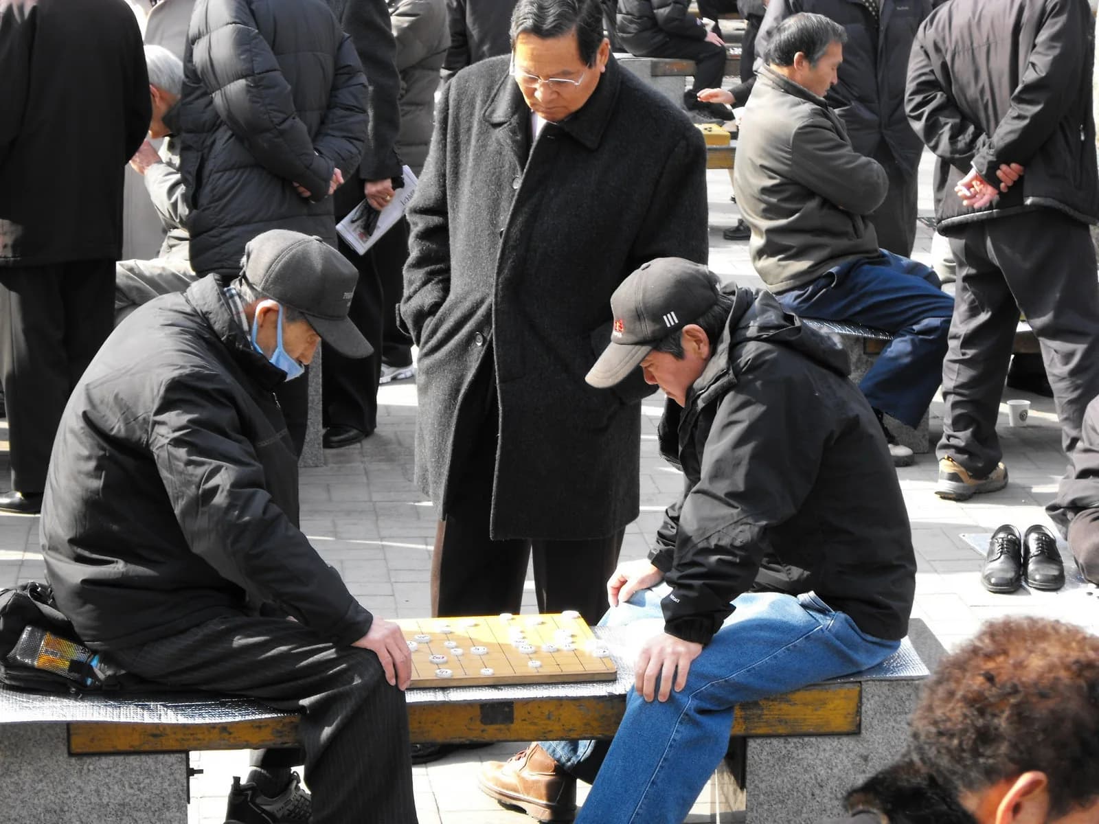 Two men play janggi on a low board in a park while several people stand nearby watching.